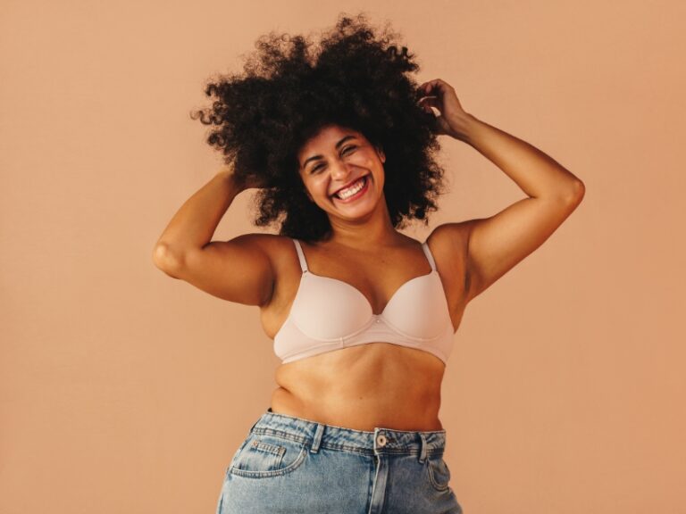 Smiling woman with curly hair wearing a bra and jeans, embracing body positivity.