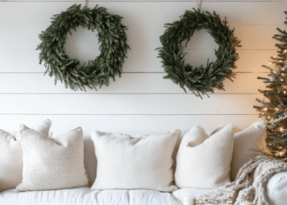 Cozy Christmas living room featuring a white shiplap wall with two decorative wreaths, a white sofa adorned with a green pine tree garland, and candles, presented in natural light with neutral colors and sharp details for aesthetic home decor photography.