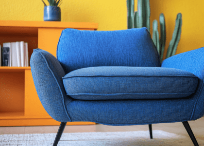 Blue fabric sofa chair with black legs in front of an orange bookcase, next to cacti on a white carpet, against yellow walls, featuring high resolution, bright colors, and soft lighting for a comfortable seating environment.