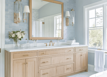 Coastal-style bathroom with a weathered oak vanity, white countertop, vintage brass hardware, soft blue ceramic tile walls and floor, large mirror, and elegant glass sconces, creating a serene atmosphere for morning routines.