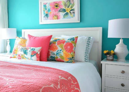 Bright and colorful bedroom featuring turquoise walls, a white headboard, white nightstand, pink pillows on the bed, vibrant wall art, and colorful floral throw blankets, creating a cheerful atmosphere.