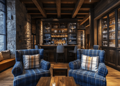 Cozy modern rustic living room featuring dark wood and blue armchairs, plaid pillows, stone walls, wooden ceiling beams, a bar, and glass-fronted cabinets beside an oak table, designed with ski resort architecture aesthetics.