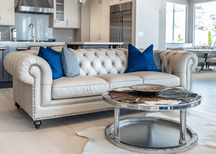 Light gray leather Chesterfield sofa with blue pillows in an open-concept living room featuring white oak flooring, a cowhide coffee table with silver legs, and a small round metal table with chrome accessories, set against a backdrop of a modern kitchen with stainless steel appliances.