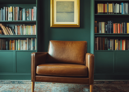 Midcentury modern leather armchair in a stylish American living room, featuring built-in shelves and bookshelves, with brown leather upholstery, wooden legs, and a gold-framed artwork above; carpeted flooring enhances the cozy ambiance, illuminated by soft natural light.