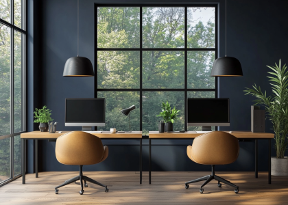 "Modern home office design featuring two computer desks with chairs, a desk lamp, large windows overlooking a forest, navy blue walls, oak wood flooring, leather armchairs, and black pendant lights above wooden tabletops in a modern farmhouse style."