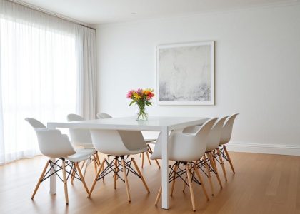 Minimalist white dining room with a sleek white table, natural wooden floor, sheer curtains allowing soft light, monochrome wall art, and a fresh flower centerpiece, creating a serene ambiance for quiet conversation and shared meals.