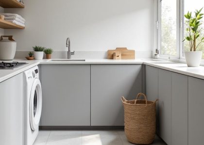 Modern minimalist laundry room with sleek gray cabinetry, smooth white countertop, tucked-away appliances, wooden shelf for towels, woven basket, and potted plants, creating a clean and warm atmosphere.