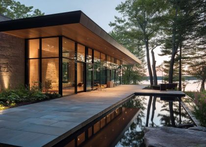 Sleek contemporary lakehouse with expansive glass walls reflecting the tranquil lake and clear sky, featuring sharp angles, a flat roof, and inviting outdoor living spaces, creating a serene ambiance harmonized with the lakeside setting.