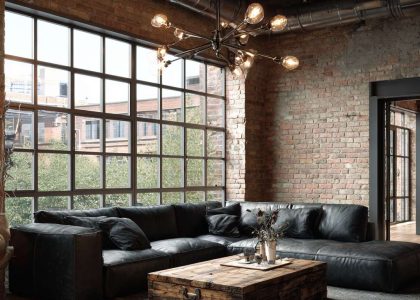 Modern industrial living room with sunlight streaming through large metal-framed windows, exposed brick walls, a low-profile black leather sofa, rustic wood coffee table, and an Edison bulb chandelier hanging from exposed ductwork, creating an inviting urban chic ambiance.