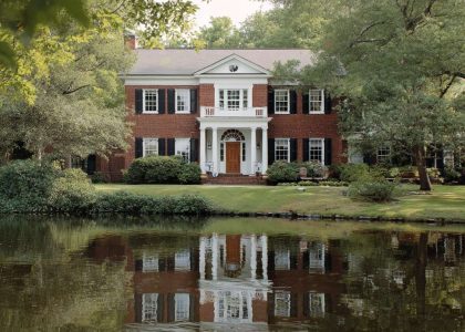 Majestic Colonial Revival lakehouse with red brick exterior, grand white portico, fluted columns, and inviting front door with fanlight, surrounded by manicured lawns and mature trees, reflecting in serene lake waters.
