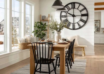 Modern farmhouse dining room with white shiplap accent wall, oversized black metal clock, natural wood accents, and sunlight filtering through large windows, creating an inviting atmosphere for family gatherings.