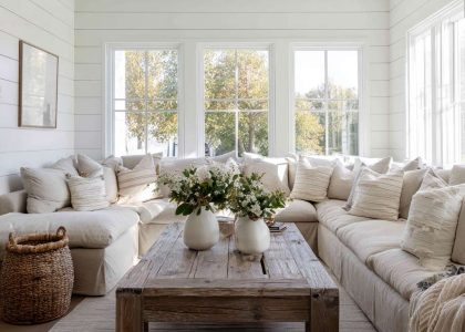 Modern farmhouse living room with large windows, white shiplap walls, oversized neutral sectional sofa with linen throw pillows, reclaimed wood coffee table, and greenery in white ceramic vases, creating a bright and inviting atmosphere.
